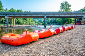 Orange float rafts with oars lined up on the bank of a river. A bridge spans across the river.
